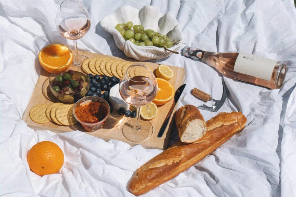 A tray of food set for a picnic with wine, grapes, biscuits, fruits, and bread sticks. 