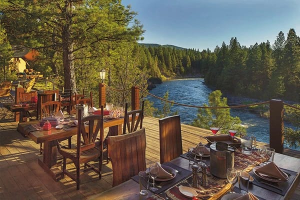 A platform with tables and chairs set for dinner overlooking a river surrounded by tall trees 