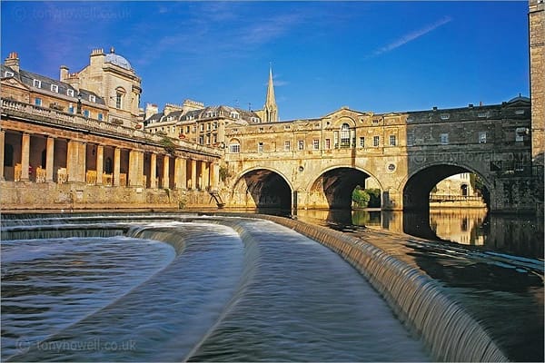 A bridge in Bath partly lit by the sun with water flowing at the bottom in neatly parted sections.
