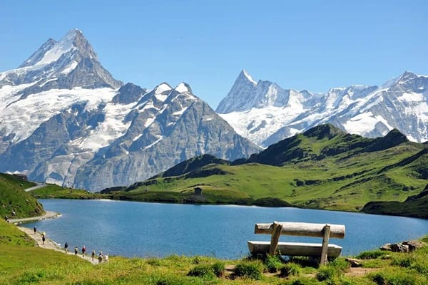 A wooden bench faced towards a river surrounded by green mountains with tall huge mountains covered in snow at the background under clear blue skies