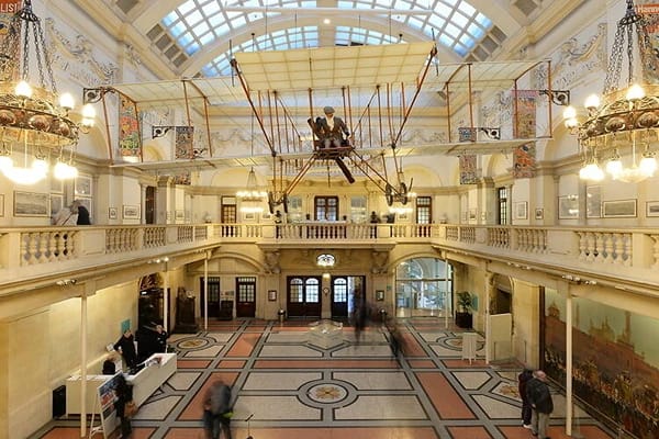 A well lit hall at Bristol Museum Art and Galley with people looking at various exhibits and a hanging aircraft artefact in the middle of the room. 