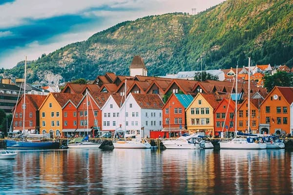 Bergen's (Bryggen) Wharf and Fjords showing deep brown coloured roofs and red or white painted houses, surrounded by mountains overlooking a body of water with boats and tourists. 