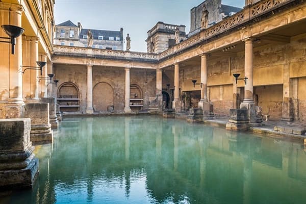 The Roman Bath in Bath with blue-green waters and old buildings around. 