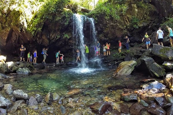 Tourists are under a waterfall with rocks and clear water in the pond.