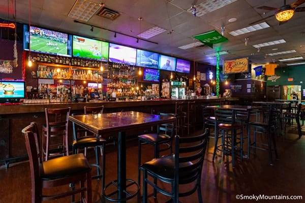 A bar with many lights and multiple televisions showing sports tables and chairs and drinks on the shelf