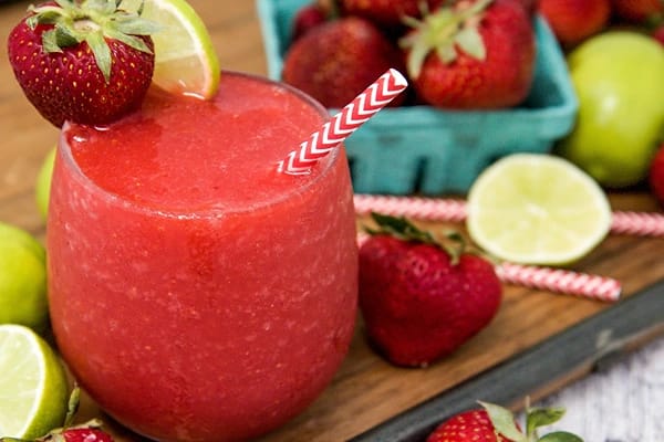 A glass of a daiquiri with a straw and a strawberry and a slice of lime fixed on it with strawberries and limes in the background.