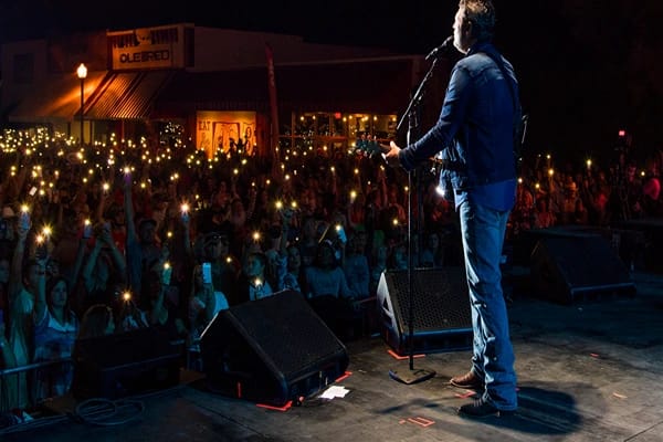 A man plays the guitar and sings in front of a crowd, raising their phone with flashlights under the night sky.