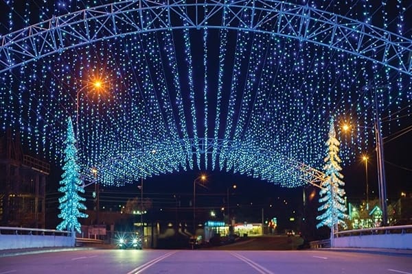 A road with many blue stringed lights hanging and two Christmas trees with blue lights at the side.