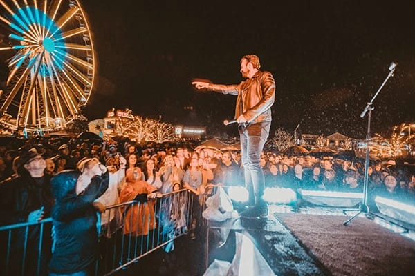 A man performing in front of a huge crowd of people at night with bright lights and a Ferris wheel in the background.