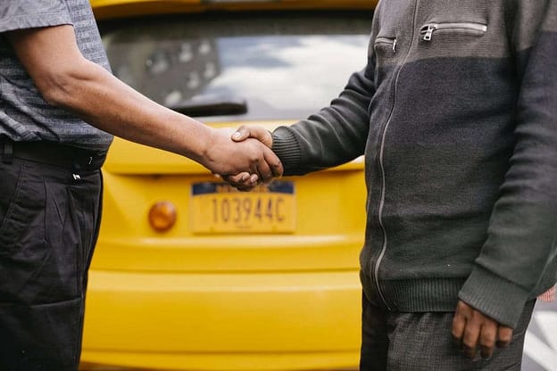 Two men. a black and white having a handshake behind a yellow painted taxi
