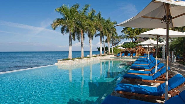 Blue seats under umbrellas in front of a long pool with palm trees. Hotel & Villas, Jamaica