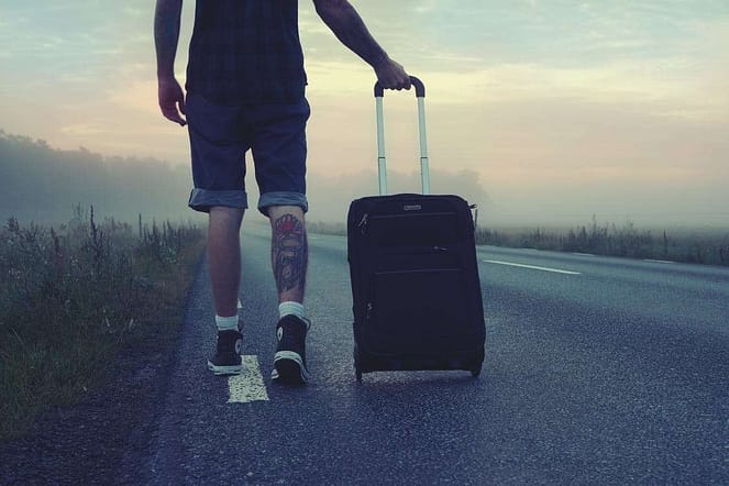 A white man with jean shorts and sneakers dragging his suit case on a lonely road.