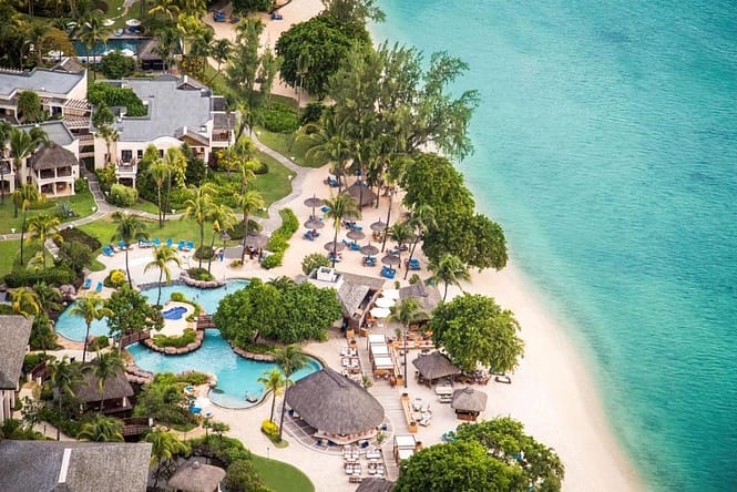 Aerial view of Hilton Mauritius Resort & Spa, a beach resort with thatched roofs, buildings, swimming pools and palm trees close to the sea shore.