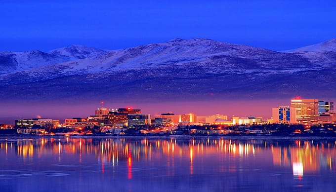 A blue night sky over a large body of water and a well lit city with large mountains in the background