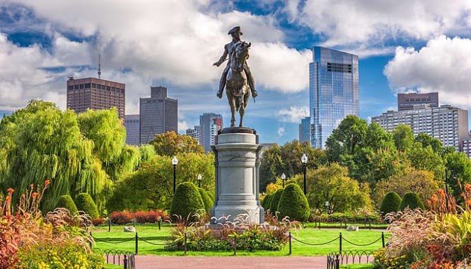A sculpture of a man riding a horse surrounded by green trees and flowers with tall buildings in the background