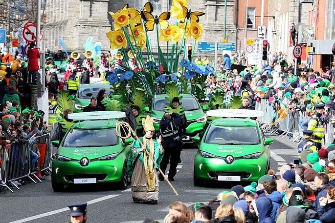 A scene from St. Patrick's Festival – Dublin, Ireland where groups of people are sitting by the roadside while green vehicles and others with costumes parade themselves