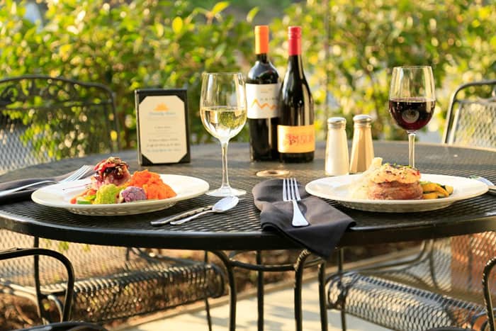 An outdoor table filled with red wines, glass cups, spoons and foods.