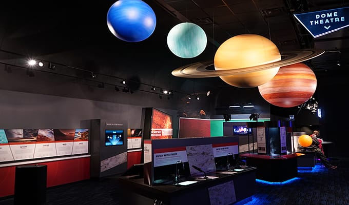A large, dark room with multiple colored displays, and some planetary bodies hung from the roof.