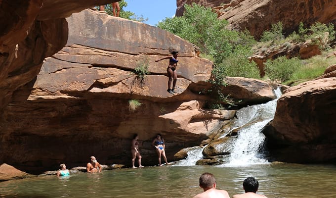 Tourists bathing in a stream and a female tourist about to jump from a waterfall into the stream